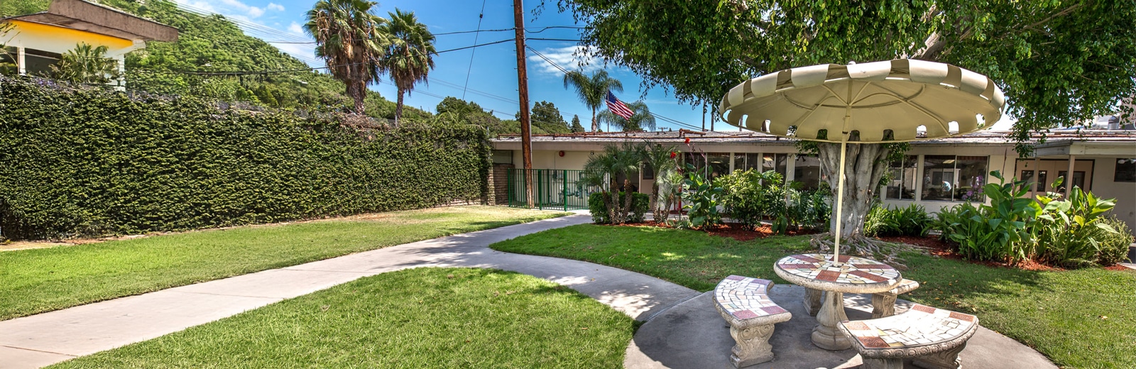 A garden features a circular stone table with matching benches under a large umbrella. It's surrounded by lush greenery and a pathway leads to a building.